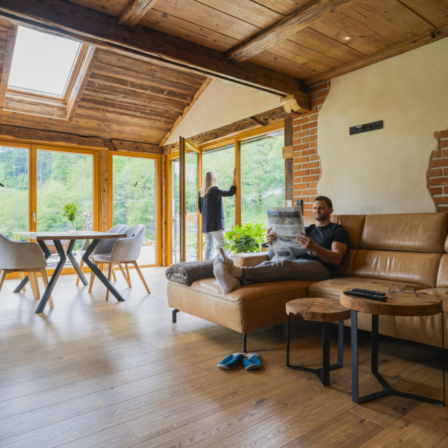 Ein junges Paar hat die Ferienwohnung AB bezogen. Er liest Zeitung auf der Couch, Sie steht in der Tür und genießt den Ausblick.
