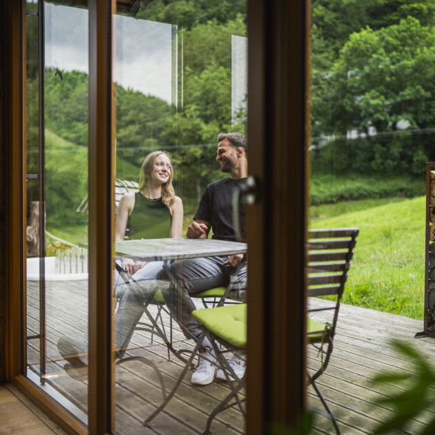 Durch die Scheibe ist ein junges Paar auf der Terrasse zu sehen. Sie sitzen am Tisch und unterhalten sich.